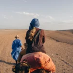 Two people riding camels through a desert landscape.