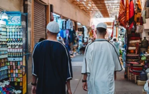 In the picture, two foreign tourists in traditional Moroccan attire walk through the narrow, cobblestone streets of old Marrakech. Their expressions show joy and amazement as they immerse themselves in the rich culture and historic charm of the city.